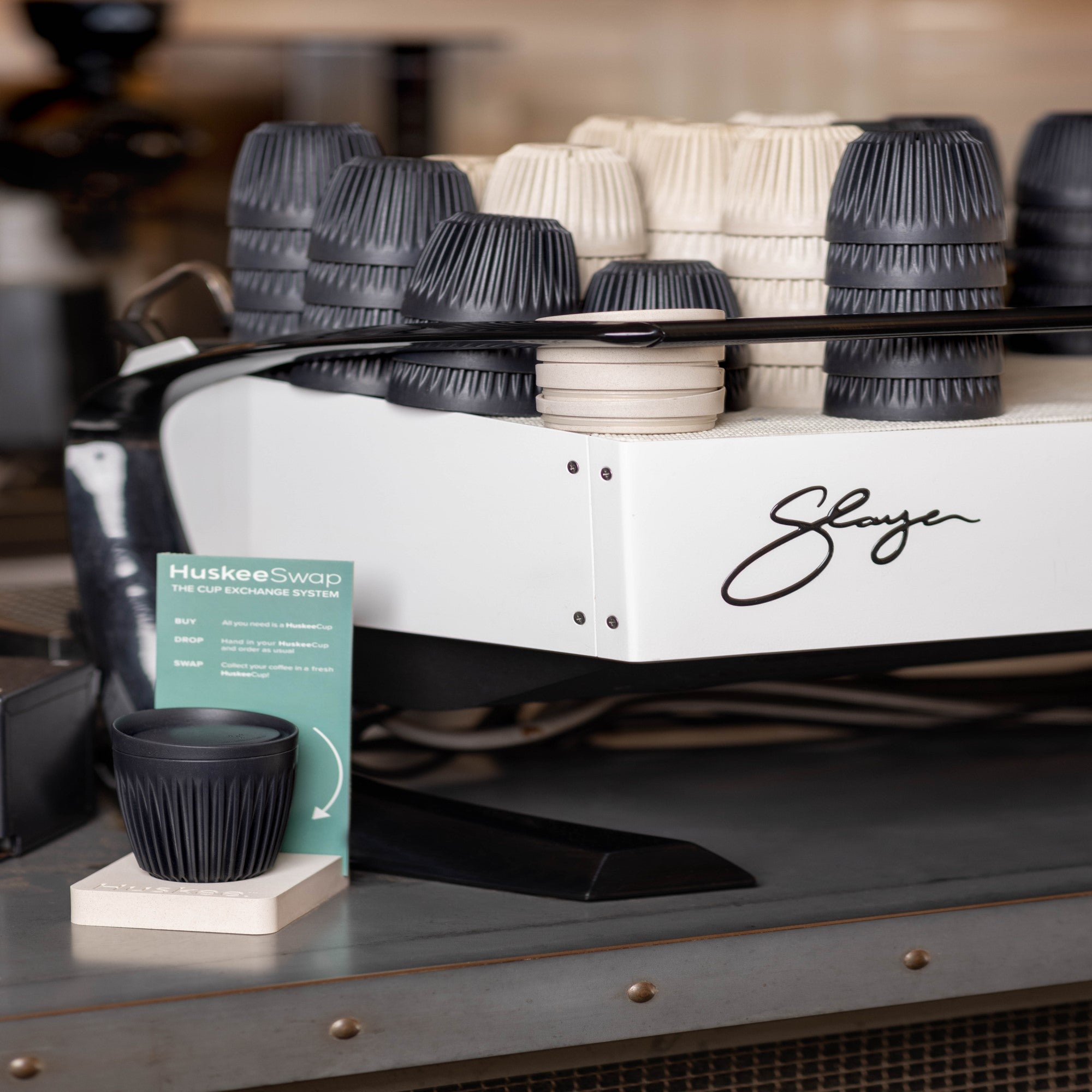 Eco-friendly coffee cups stacked on a coffee machine with HuskeeSwap exchange system sign